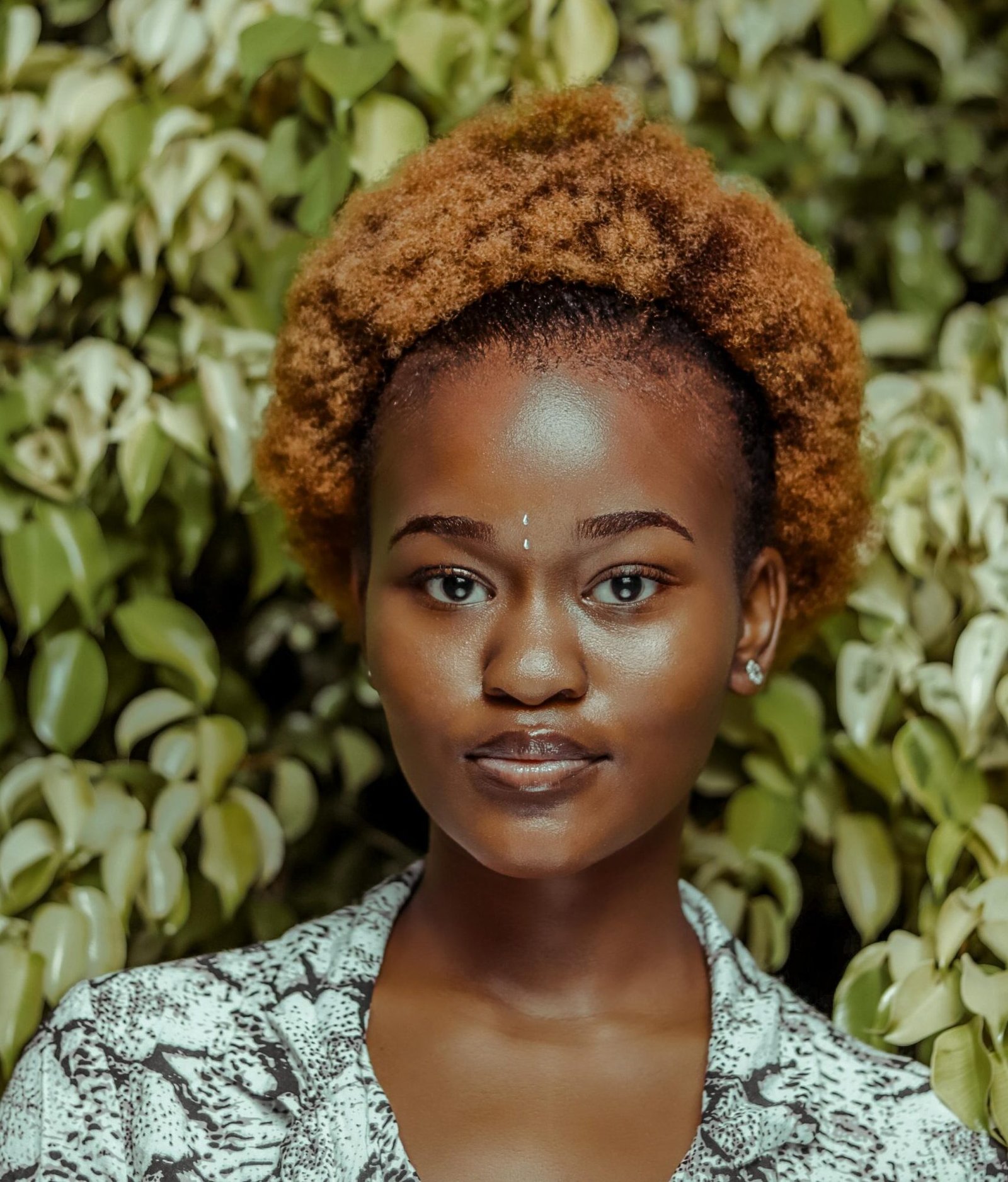 Portrait of a young woman with an afro hairstyle in front of vibrant green foliage.